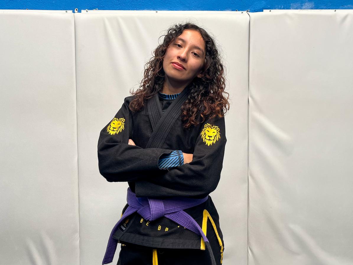 Karen guerra posando de brazos cruzados en atavío de Jiu Jitsu contra una pared acolchada blanca